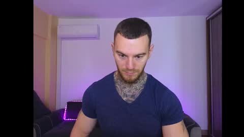 Media: Video of a muscular, bearded man with short dark hair wearing a black V-neck t-shirt and a patterned bandana. Background shows a beige wall with an air conditioner and purple LED lights.