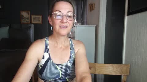 Media: Video of a middle-aged Caucasian woman with glasses, wearing a blue and white patterned sports bra, sitting in a wooden chair, in a dimly lit room with framed pictures on the wall.
