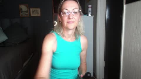 Media: A video of a middle-aged woman with shoulder-length blonde hair, wearing glasses and a turquoise tank top, standing in a dimly lit bedroom.
