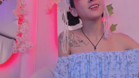 Media: Video of a smiling Asian woman with fair skin, wearing a blue floral off-shoulder top, intricate floral tattoo on her right shoulder, and white floral garland in the background.