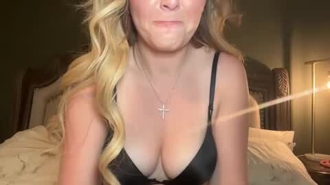 Media: Video of a blonde woman with fair skin, wearing a black lace bra, and a silver cross necklace, posing indoors, with dim lighting.