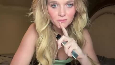 Media: A video of a blonde, fair-skinned woman with long, wavy hair, wearing a green top, using a black mascara wand to cover her blue eyes. She is indoors, likely in a bedroom, with a neutral-colored background.