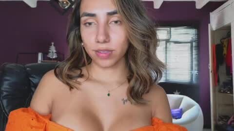 Media: Video of a young woman with light brown skin, shoulder-length wavy hair, and a small tattoo above her collarbone. She's wearing an orange off-shoulder top, sitting in a purple room with a black leather chair, a window with blinds, and a white chair.