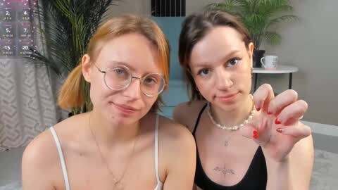 Media: Video of two young Caucasian women with fair skin, one with red hair and glasses, the other with dark hair and a pearl necklace, both smiling in a cozy room with greenery and a patterned curtain.