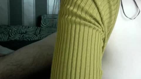 Media: A close-up video shows a person's lower leg and torso in a mustard-yellow ribbed knit sweater, with a striped sofa and patterned wall in the background.