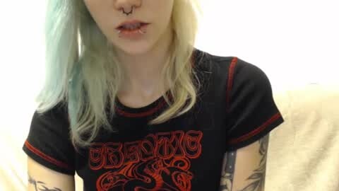 Media: Video of a pale-skinned woman with shoulder-length, pastel blue hair, wearing a black Slayer t-shirt with red lettering, and a septum piercing. She has multiple tattoos on her arms, and is sitting on a cream-colored couch.