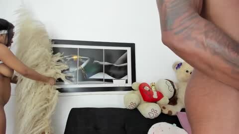 Media: Video of a naked woman with dark skin and medium-sized breasts, holding a feather duster, in a modern room with a black sofa, plush toys, and abstract art on the white wall.