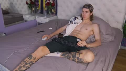 Media: Video of a shirtless, tattooed man with long hair, wearing black shorts, sitting on a bed playing a video game, in a modern bedroom.