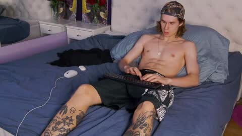 Media: Video of a shirtless, tattooed man with long hair in camo hat, black shorts, and blue sheets, typing on a laptop in a modern bedroom.