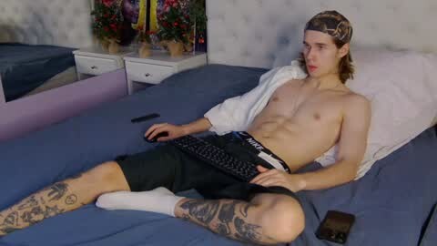 Media: Video of a shirtless, lean white man with long brown hair, wearing black shorts, sitting on a bed, holding a keyboard.
