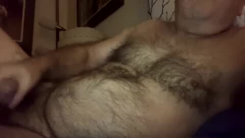 Media: Video of a partially nude, overweight man with a hairy chest, lying on a bed. His right hand is pulling down his pants, revealing a large, uncircumcised penis. The background is dimly lit with a beige wall and a dark, blurry object.