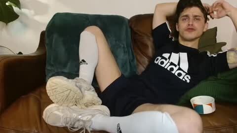 Media: Video of a young, fit man with short dark hair, wearing a black Adidas athletic shirt and shorts, lying on a brown leather couch. He has white knee-high socks and white sneakers.
