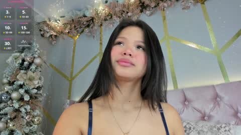 Video of a young woman with straight black hair, light skin, and a slender physique, wearing a blue spaghetti strap top. She stands indoors with a festive background featuring a pink tufted headboard, snow-covered Christmas tree, and gold-framed walls.