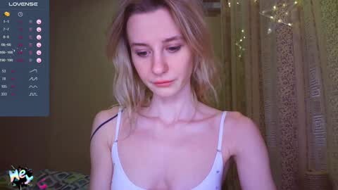 Media: A video of a young, slender, fair-skinned woman with long blonde hair, wearing a white tank top, looking down, in a dimly lit room with a TV displaying \"LOVENBIZ\" and social media icons.