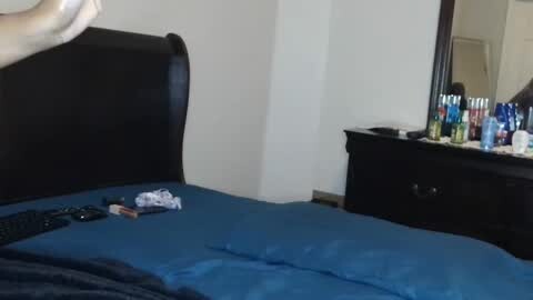 Media: A video of a bedroom with a black upholstered bed and blue bedspread. A black dresser with a mirror and numerous bottles of alcohol reflects a person partially visible in the doorway.