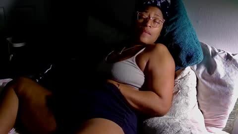 Media: A video of a plus-sized, middle-aged Black woman with glasses, wearing a grey sports bra and navy shorts, lying on a bed with patterned pillows, looking relaxed.