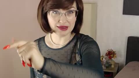 A video of a middle-aged woman with a short, dark bob haircut, glasses, and red lipstick, wearing a sheer black top, pearls, and red nails, gesturing with her hands, indoors with a beige wall and a dark headboard in the background.