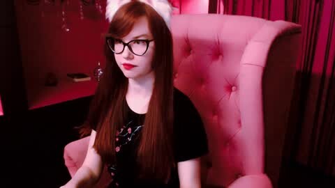 Media: Video of a pale-skinned woman with long brown hair and glasses, wearing a black floral shirt, white headband, and red lipstick, sitting in a pink tufted chair under pink lighting in a dimly lit room.