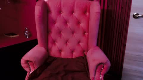 Media: Video of a plush, pink, tufted armchair with nailhead trim, set against a vibrant red wall and a partially visible white door in a modern, minimalist room.