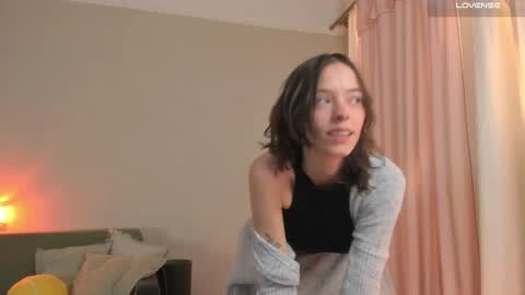 Media: Video of a young woman with shoulder-length dark hair, wearing a black top and gray cardigan, leaning on a sofa, with beige walls, a green couch, and a lit lamp in the background.