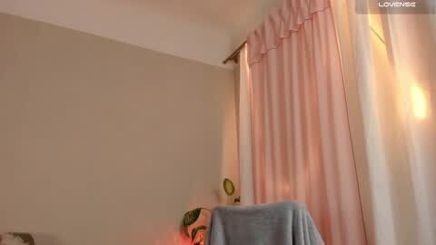 Media: A video of a simple, beige-walled bedroom with a pink curtain and a teal blanket draped over a chair, bathed in warm, soft lighting.