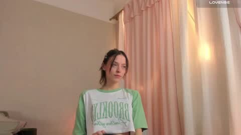 Media: A young woman with fair skin and dark hair, wearing a green and white t-shirt, stands in a dimly lit room with soft pink curtains and a beige wall.