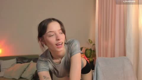 Media: A video of a young, slim, fair-skinned woman with dark hair, wearing a grey shirt and black panties, leaning on a couch with a potted plant in the background.