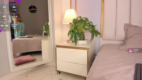 Media: Video of a cozy bedroom with a beige bed, white nightstand, green plant, pink throw pillow, and a lamp, with a person sitting on the bed in a teal dress.