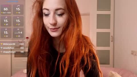 Media: Video of a young woman with long, vibrant red hair and fair skin, smiling in a bedroom with a pink bedspread and a digital thermometer on the wall.