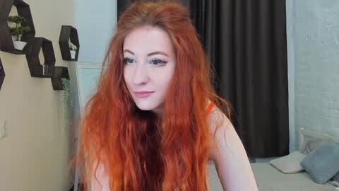 Media: Video of a pale-skinned woman with long, vibrant red hair, wearing a sleeveless top, sitting in a room with black curtains, beige walls, and a textured white brick wall.