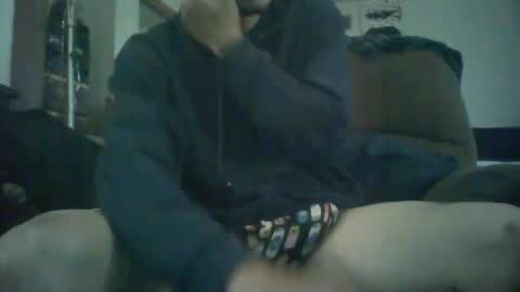 Media: A blurry video shows a person sitting on a couch, wearing a dark hoodie and colorful shorts, with a hand near their mouth, in a dimly lit room.