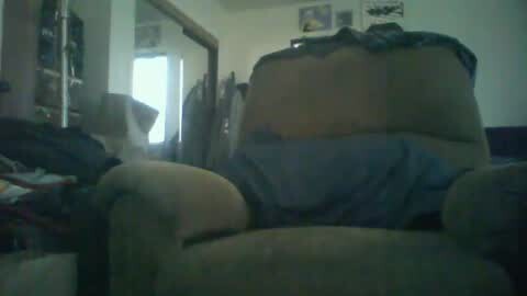 Media: A dimly lit video of a cluttered living room with a beige armchair draped in a blue blanket. Clothes and clutter cover the floor and furniture.