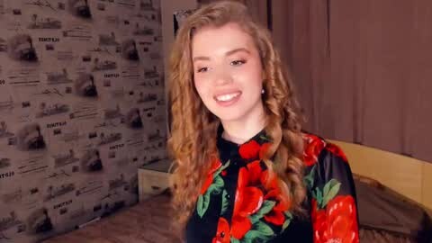 Media: Video of a young woman with curly blonde hair, smiling, wearing a black top with large red and green floral print, in a room with gray newspaper-patterned wallpaper and a bed.