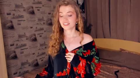 Media: Video of a smiling young woman with long, curly blonde hair, wearing a black floral dress, standing in a room with newspaper-patterned wallpaper and a yellow couch.