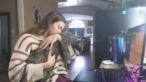 Media: Video of a young woman with long brown hair, wearing a striped sweater, kissing a large brown dog. Background shows a modern living room with a kitchen, bookshelves, and a computer monitor.