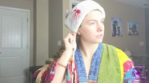 Media: Video of a woman with a white headwrap featuring red and green embroidery, wearing a colorful, patterned robe with a blue vest, in a beige-toned room with framed artwork and a closed door.