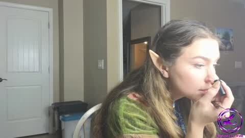 Media: Video of a young girl with long brown hair, wearing a green t-shirt, and elf ears, sitting at a table, talking on a phone, in a modern beige room with a closed door, a light switch, and a framed picture on the wall.