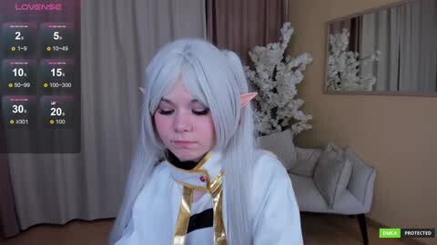 Media: Video of a young woman with long silver hair and elf ears, wearing a white cloak with gold accents, in a modern living room with a white floral arrangement.