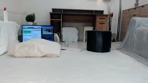 Media: Video of a partially furnished living room with white walls, a dark wood desk, and a computer screen displaying a weather forecast. A beige cushion, a black cylinder, and a white chair are visible.