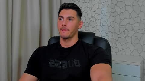 Media: Video of a muscular, light-skinned man with short dark hair, wearing a black \"GAINS\" t-shirt, seated in a black chair against a textured white wall.