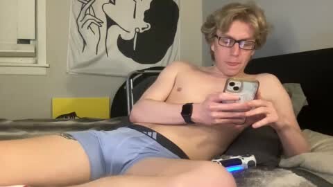 Video of a shirtless, slim, light-skinned man with glasses and short curly hair, wearing blue shorts, lying on a bed, holding a smartphone. Background includes a black and white poster and gaming console.