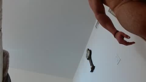 Media: Video of a bare-chested man with a muscular build, standing in a white-walled room, with a wall-mounted black electrical outlet and a red cord visible.