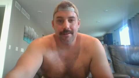 Media: Video of a middle-aged, shirtless man with a beard and light hair wearing a backward baseball cap, smiling. He's in a modern living room with beige walls, a TV, and a brown couch.