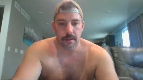 Media: Video of a shirtless, light-skinned, bearded man with a tan line from a baseball cap, indoors near a couch with a window and gray curtains.
