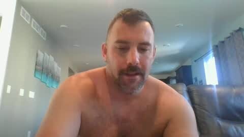 Media: Video of a shirtless, muscular white man with short dark hair and a beard, slightly smiling, in a modern living room with gray furniture and light blue curtains.