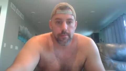 Media: Video of a shirtless, middle-aged white man with a beard, wearing a backward baseball cap, sitting indoors. The background features a modern living room with light-colored walls and a large window.