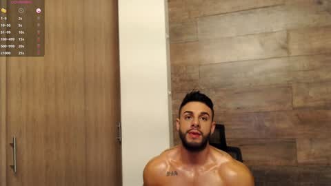 Media: Video of a muscular, shirtless man with short black hair and a trimmed beard, sitting in a modern bathroom with wooden walls and a calendar display in the top left corner.
