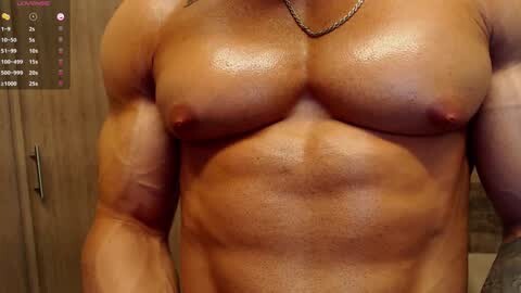 Media: Video of a shirtless, muscular man with tanned skin, showing off defined pectorals and abs, wearing a thin chain necklace. Background features a wooden door and a calendar with dates.