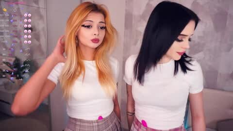 Media: Video of two young women with long, straight hair, one blonde and the other black. They wear tight white crop tops and plaid skirts, standing side-by-side in a modern, softly lit room.