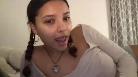 Media: Video of a young woman with medium brown skin, dark braided hair, wearing a beige long-sleeve top, silver hoop earrings, and a heart-shaped pendant necklace, posing indoors with a light-colored couch and patterned curtains in the background.